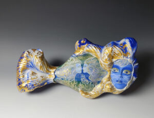 Ceramic fish shaped sculpture. The predominate colors are blue and gold. The fish has the face of a person and has a line drawing of two silhouettes on the stomach area.