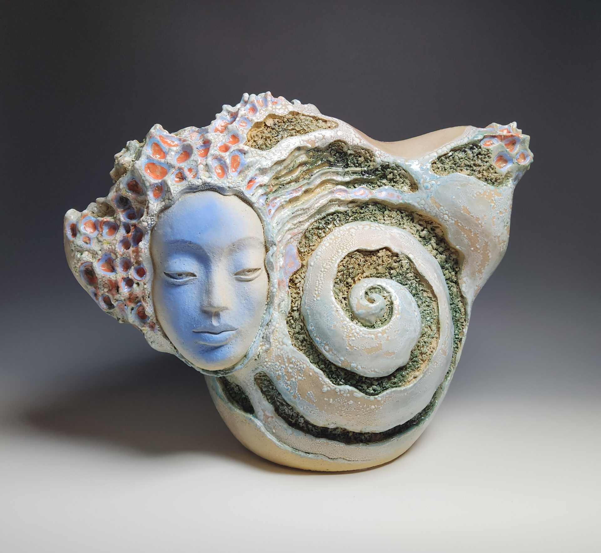 Ceramic sculpture. The form slightly resembles a shell and has a spiral in the middle. There is a blue face with flowing hair protruding from the overall shell shape.