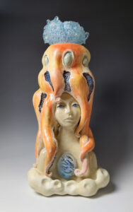 This is a tall ceramic sculpture. It is a bust of a female with what seems to be an octopus for hair. The head of the octopus seems to be mad out of blue glass.